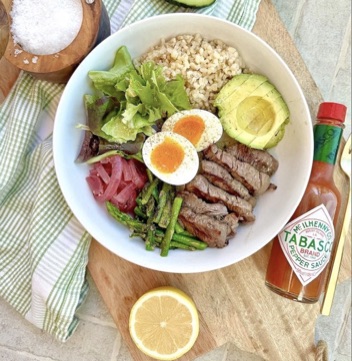 Lunch bowl with sliced steak, soft-boiled egg, avocado, asparagus, brown rice, mixed greens, and pickled red onion, with a bottle of Tabasco and a lemon wedge on the side